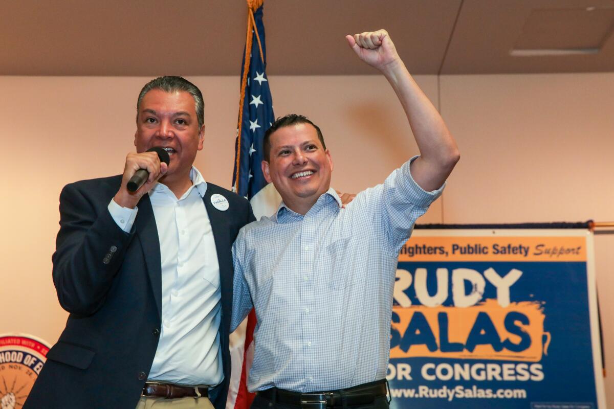 One man speaks into a microphone next to a man raising his fist