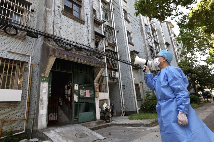 Un uomo mascherato che indossa guanti e un camice medico blu e un berretto tiene un megafono bianco fuori da un edificio 