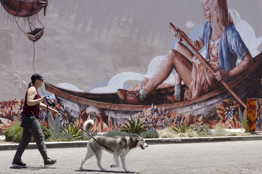 CULVER CITY, CA - JUNE 14, 2021 - Jen and her dog Biscuit walk past a mural by Australian artist Fintan Magee where no water is in sight for a rower on a very hot afternoon along Lindblade Street in Culver City on June 14, 20212. The mural by Magee is entitled, "The Zeppelin." The Southland finds itself baking in summer-like heat this week, with triple-digit record temperatures anticipated in the valleys and mountains, while even downtown could reach up to 100. (Genaro Molina / Los Angeles Times) FYI EDITORS: THE SUBJECT ONLY WANTED HER FIRST NAME USED.