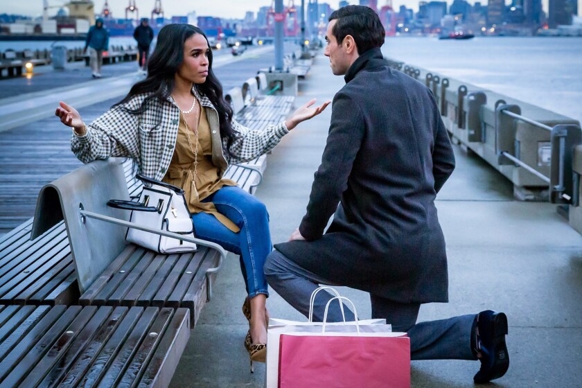 A man kneels before a woman sitting on a bench at the waterfront.