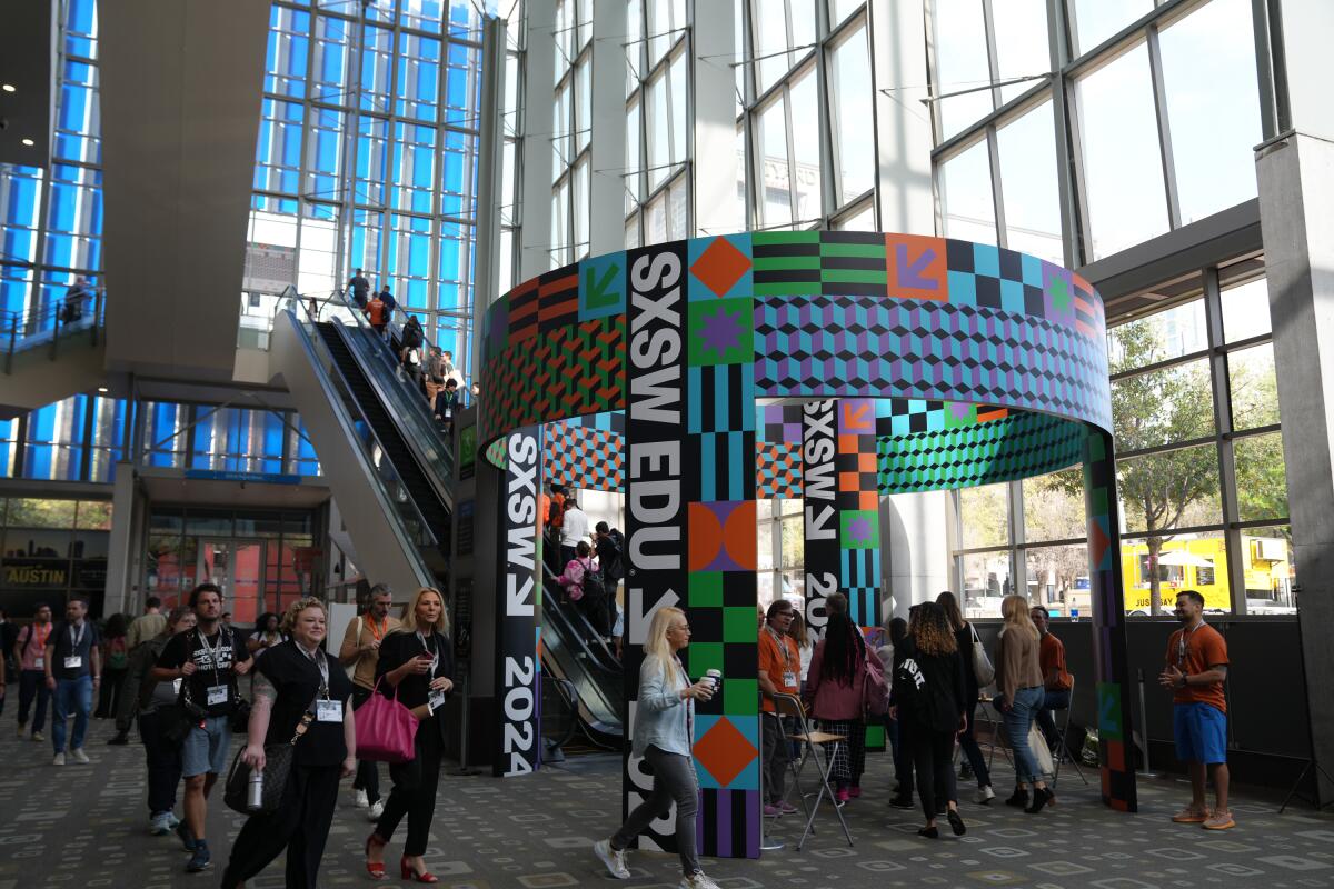 The interior of a convention center decorated with vibrant banners. Dozens of attendees stand around and use and escalator