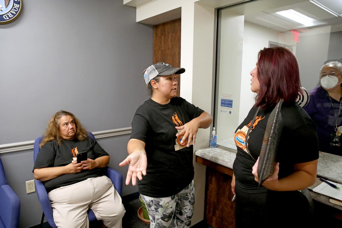 Two women talk while standing in an office as another woman sits and looks at them. 