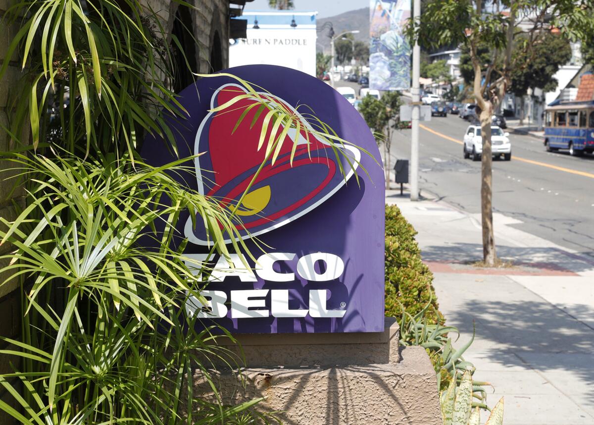 Taco Bell logo and sign stands on display on a side of a building, partially obstructed by leaves. 