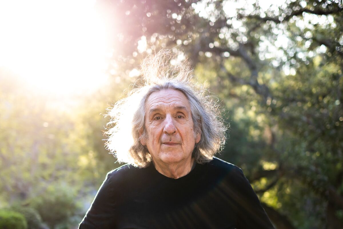 An older man in a dark shirt and with long gray hair is photographed with trees in the background.