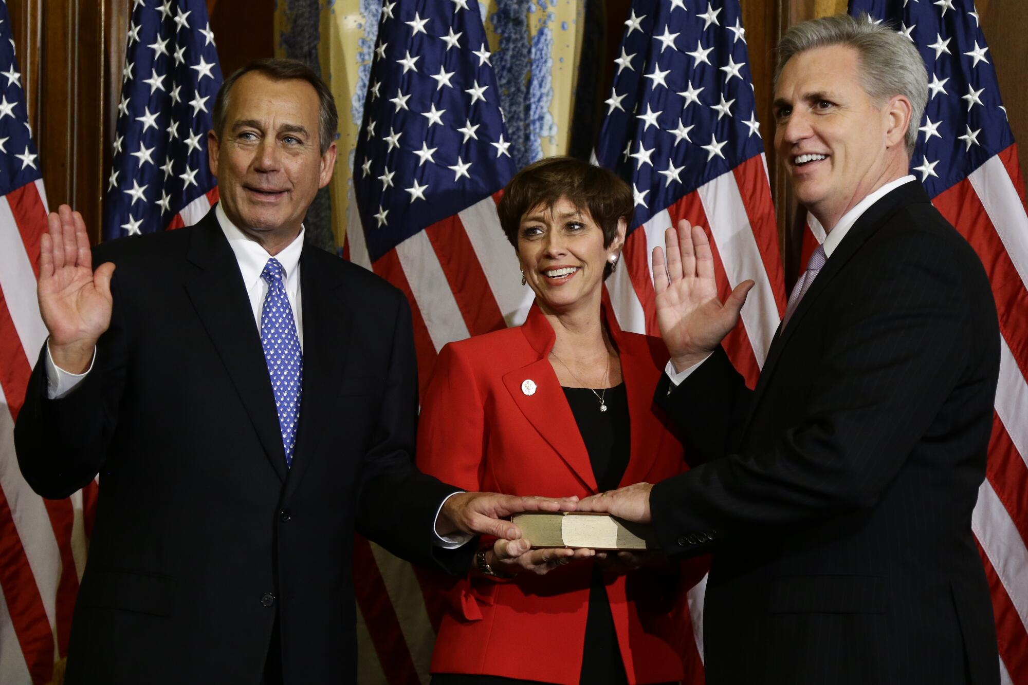 Two men in suits each hold up a hand and rest the other hand on a book held by a woman between them in front of flags.