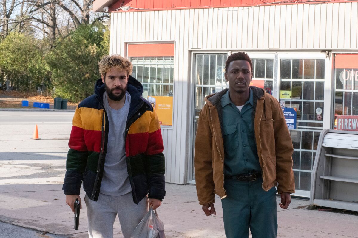 Two men in jackets stand in front of a business, one holding a gun and a sack of food.