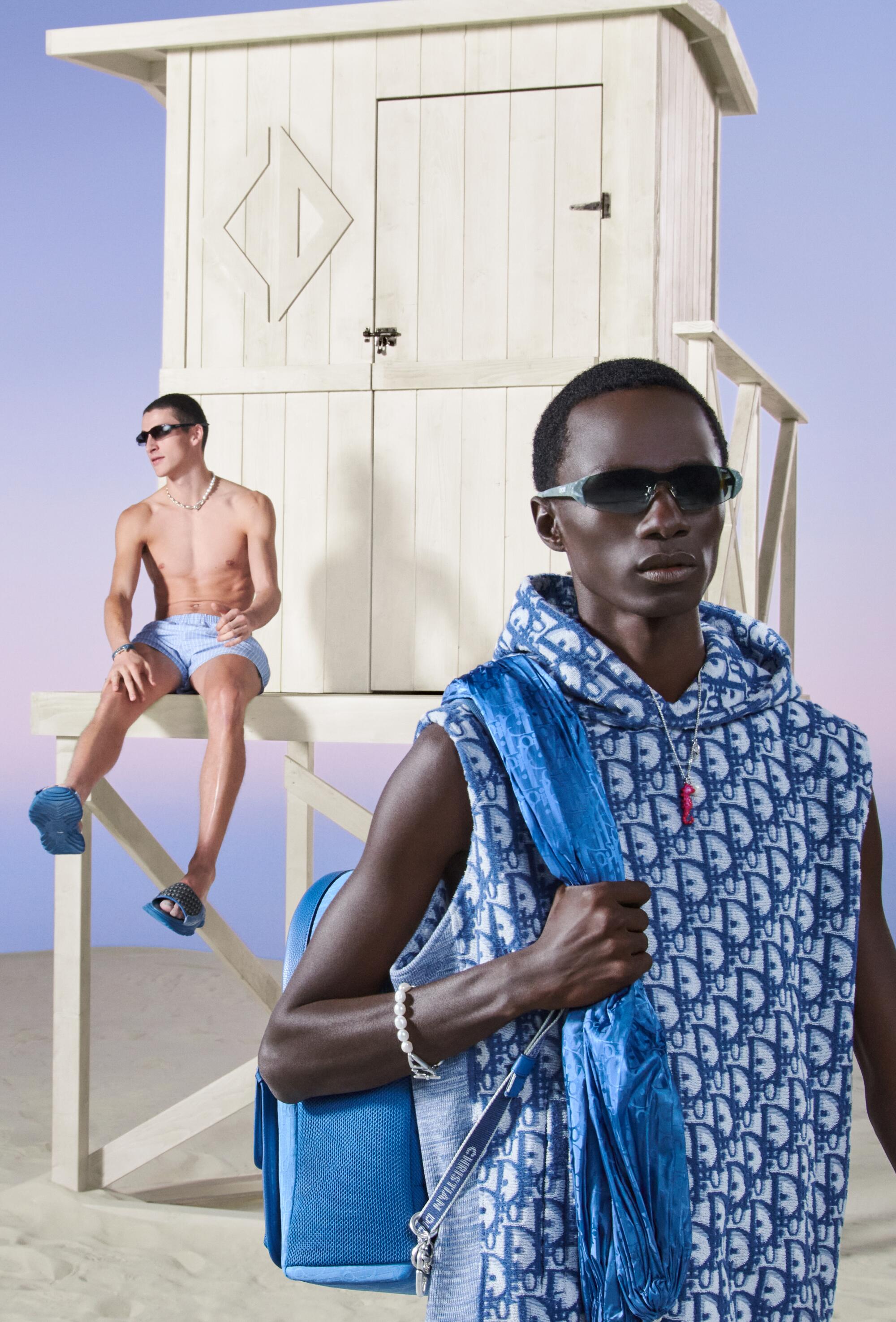 A model in shorts sitting on a lifeguard tower;  in the foreground, a model wearing a blue sleeveless hoodie and a blue towel