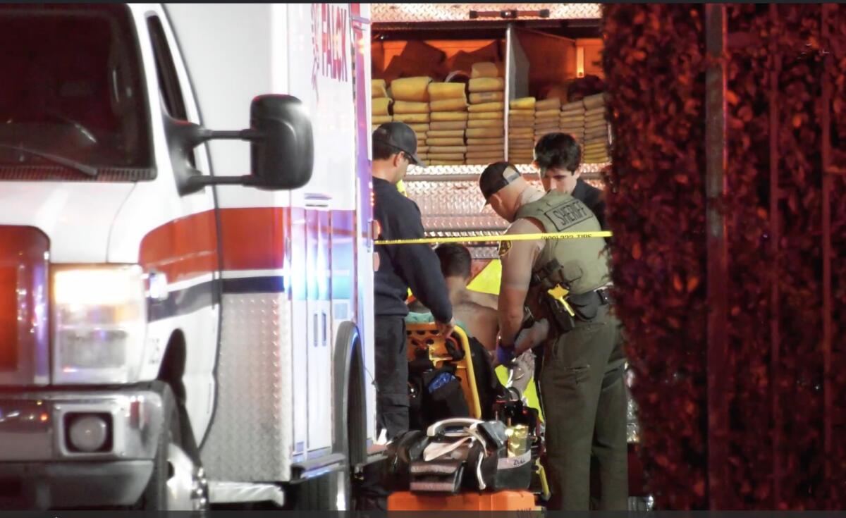 People stand around a stretcher behind an ambulance. One person is sitting on the stretcher.