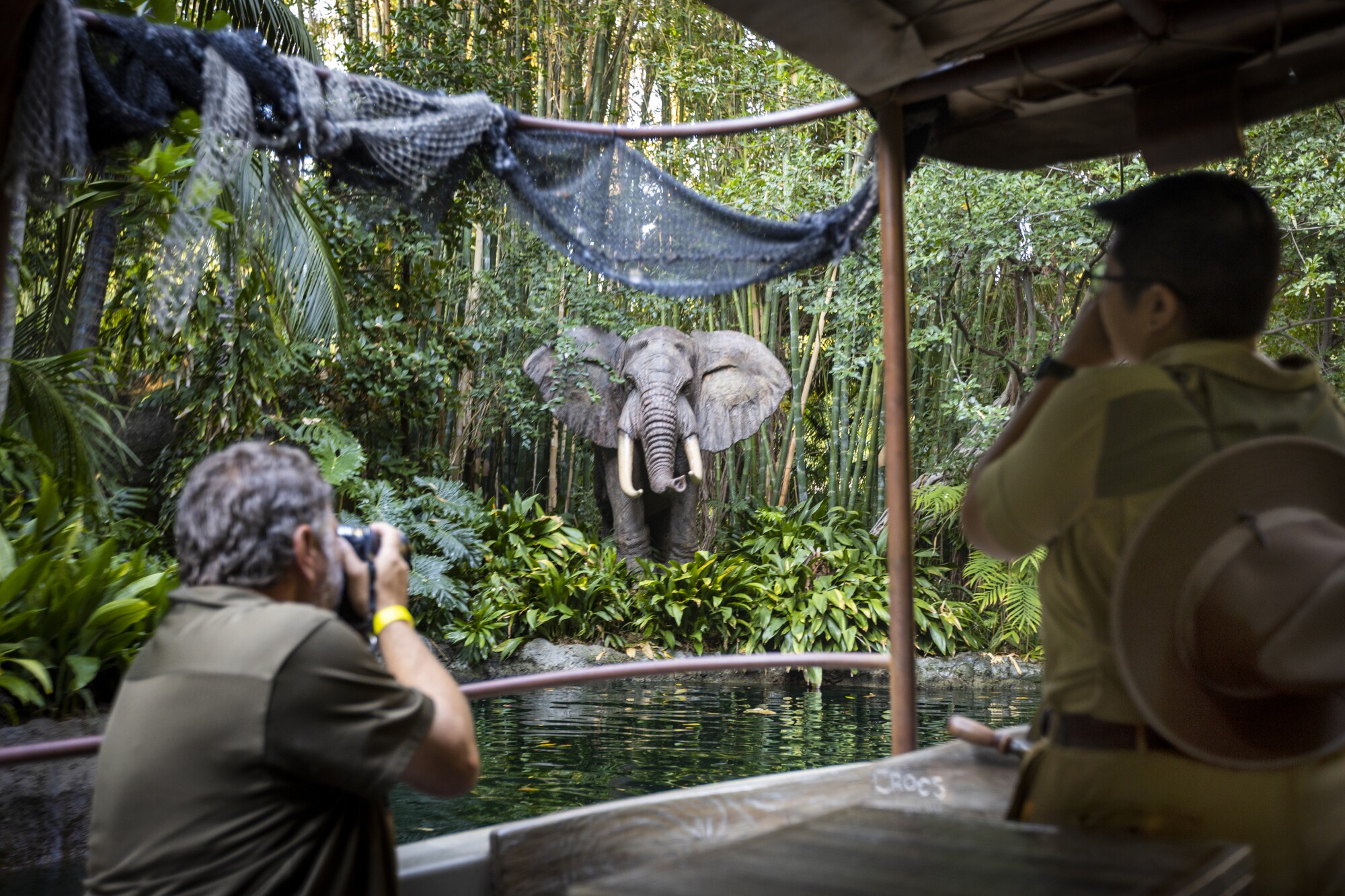 Two people look out from the Disneyland Jungle Cruise at an animatronic elephant surrounded by jungle foliage. 