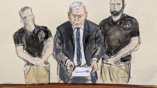 A court drawing of a man reading from a paper in a business suit with U.S. Marshals on either side