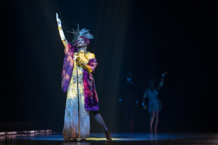 A woman in old Hollywood glam stands on a stage in multicolored light and raises her hand to the sky