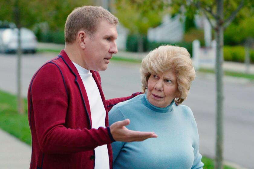 Todd Chrisley in a red cardigan holding his right hand out and standing next to his mother, Faye, in a blue turtleneck
