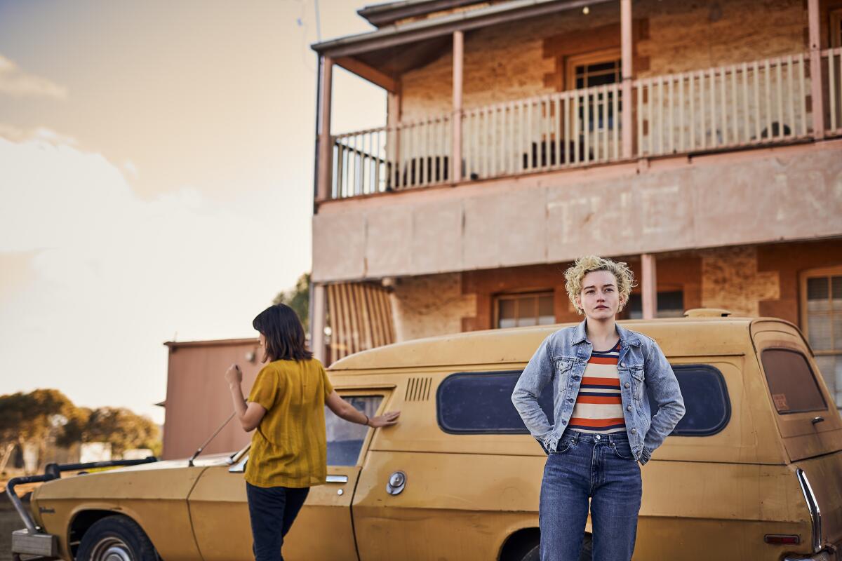 Two women stand outside a remote hotel next to a dusty old car.