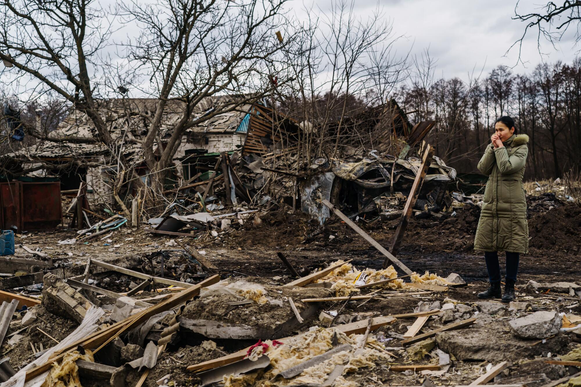 A woman clasps her hands in front of her face as she stands amid rubble.