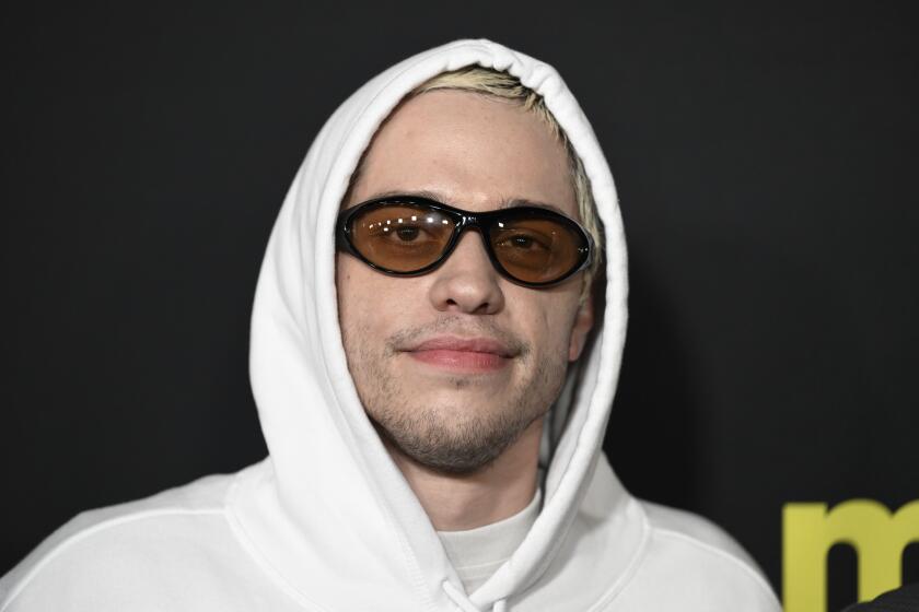 Pete Davidson wearing a white hoodie and black frame sunglasses with amber lenses, giving a half smile