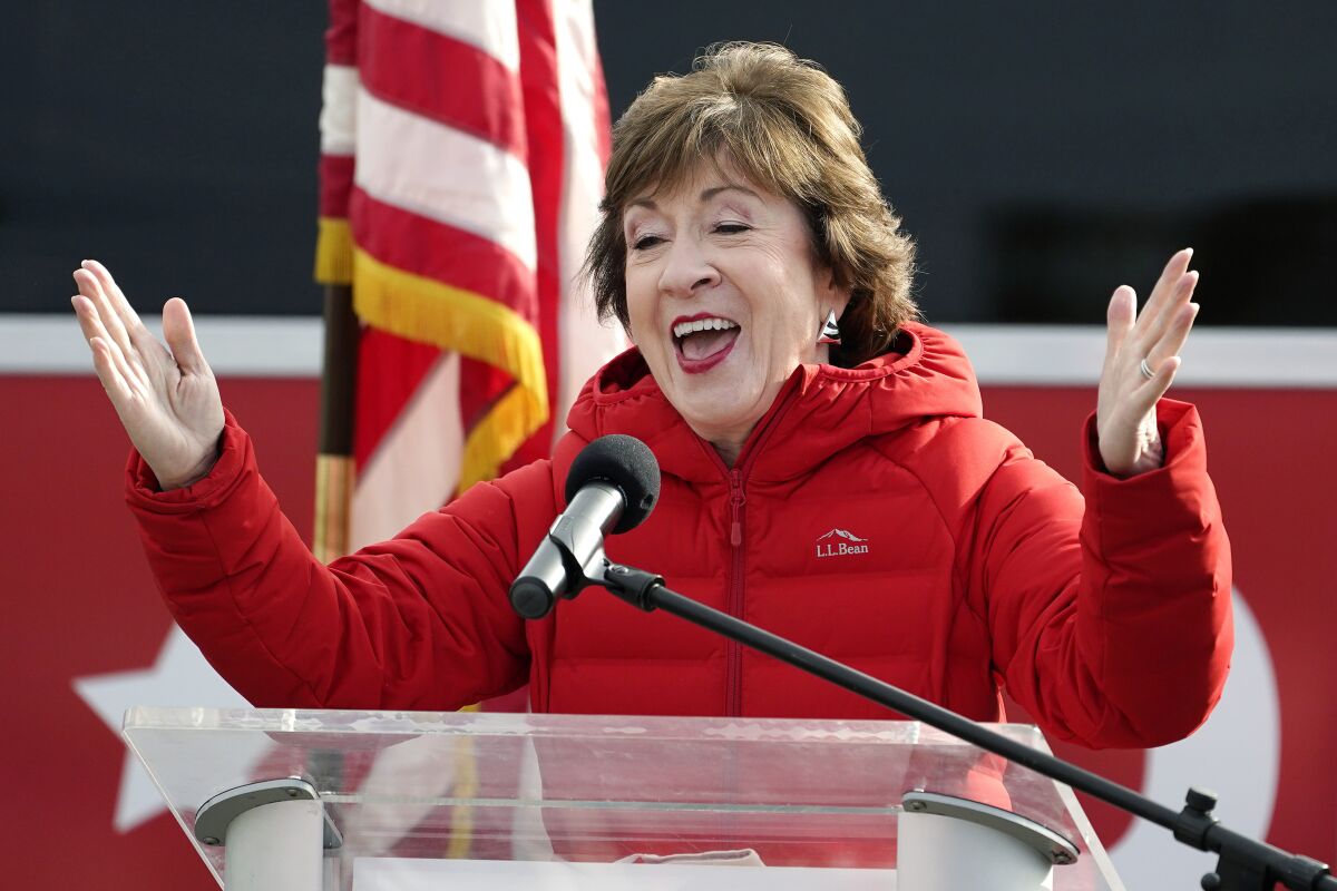 Senator Susan Collins in a red down jacket gestures with both hands in excitement while speaking at a lectern