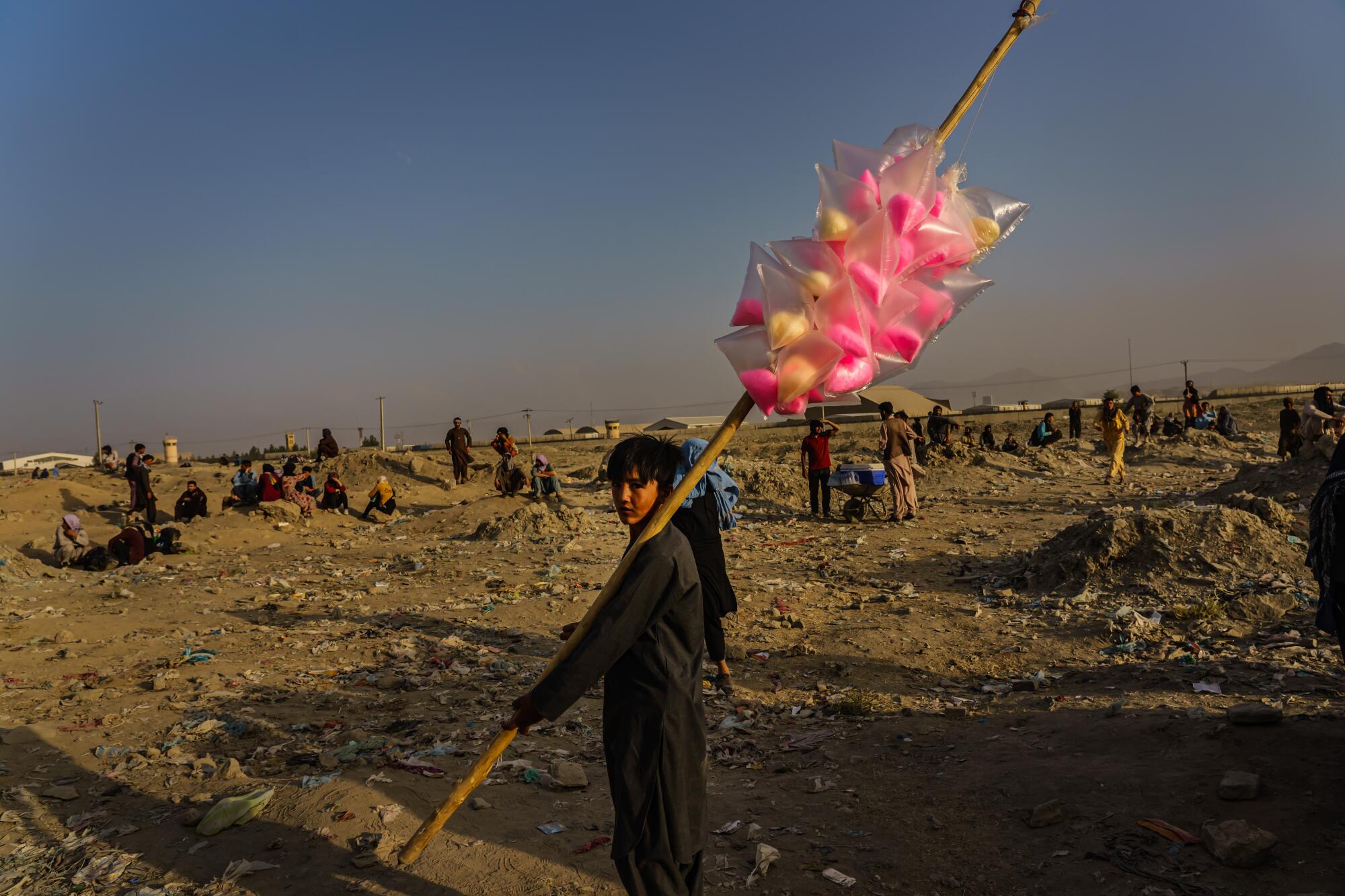 A boy holds a pole with cotton candy in an area with people standing and sitting on the ground.