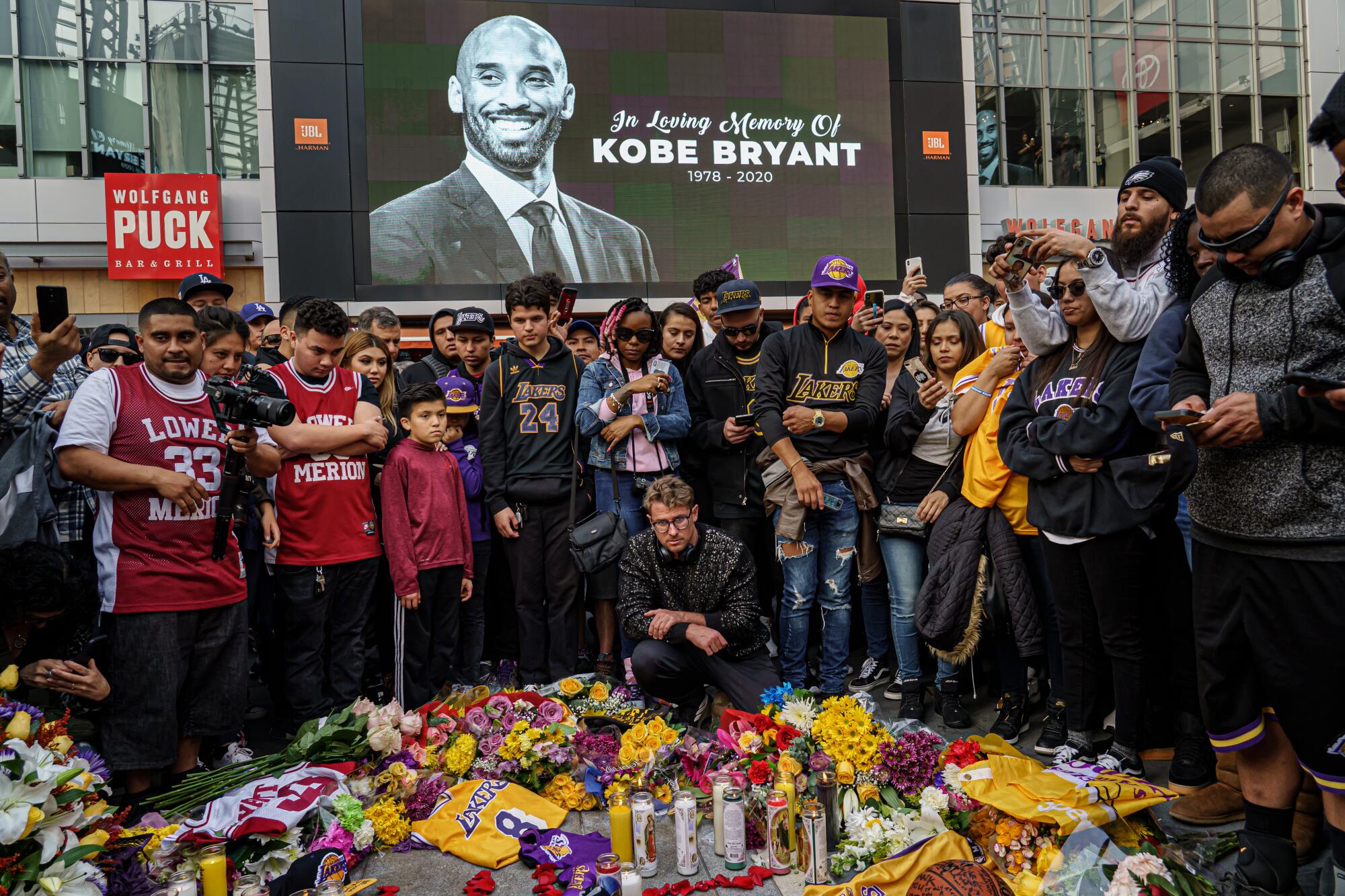 Fans gather in front of a screen with Kobe Bryant's image. On the ground is a memorial with candles, flowers and jerseys.