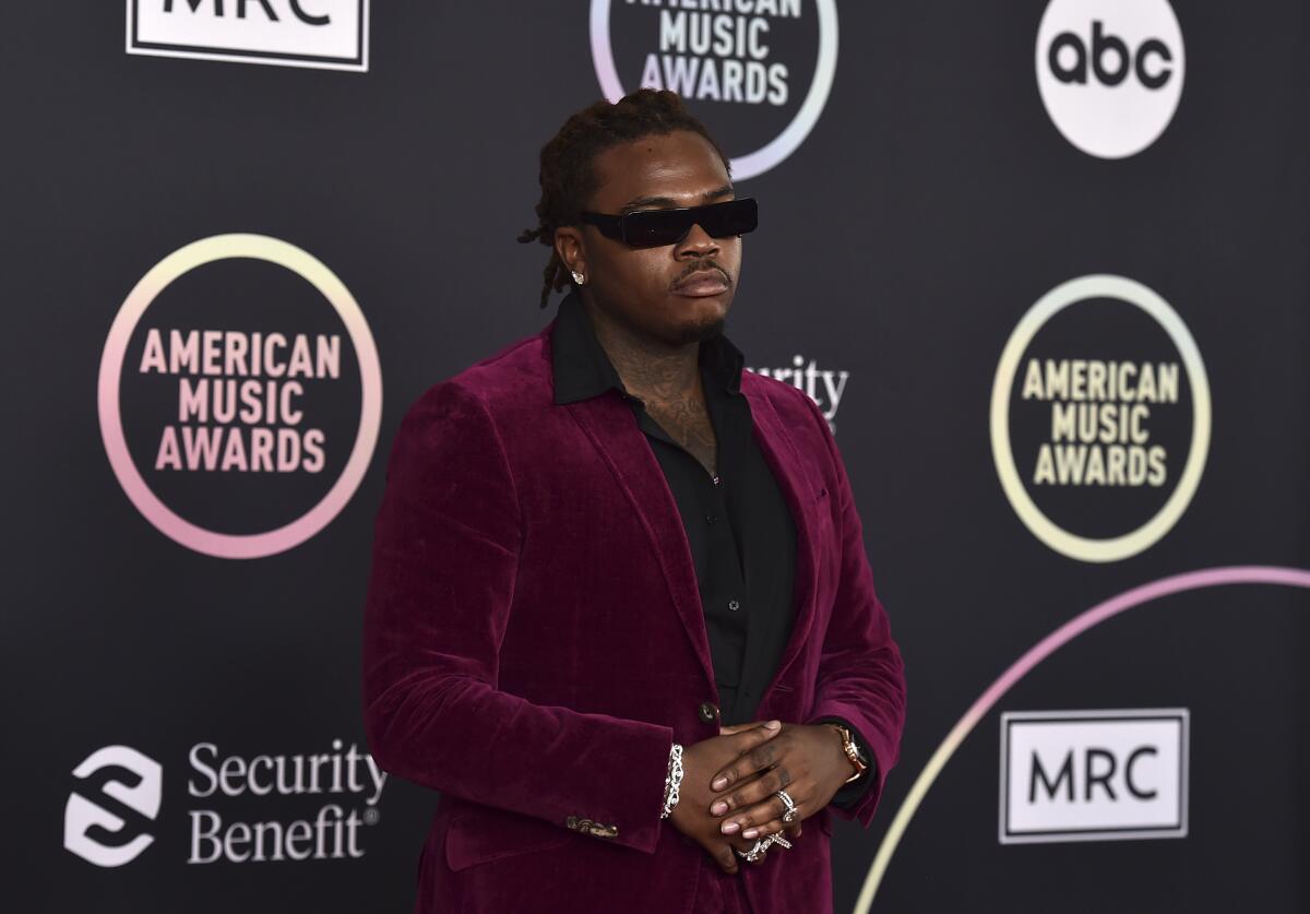 A rapper in a velvet burgundy sports coat and sunglasses walks the red carpet at an awards show.