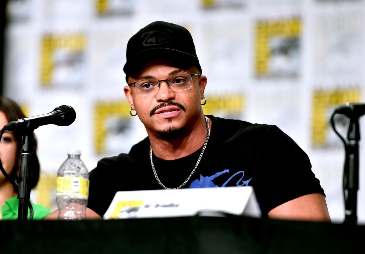 Beau DeMayo in a black T-shirt and earrings sitting at a table for a Comic-Con panel