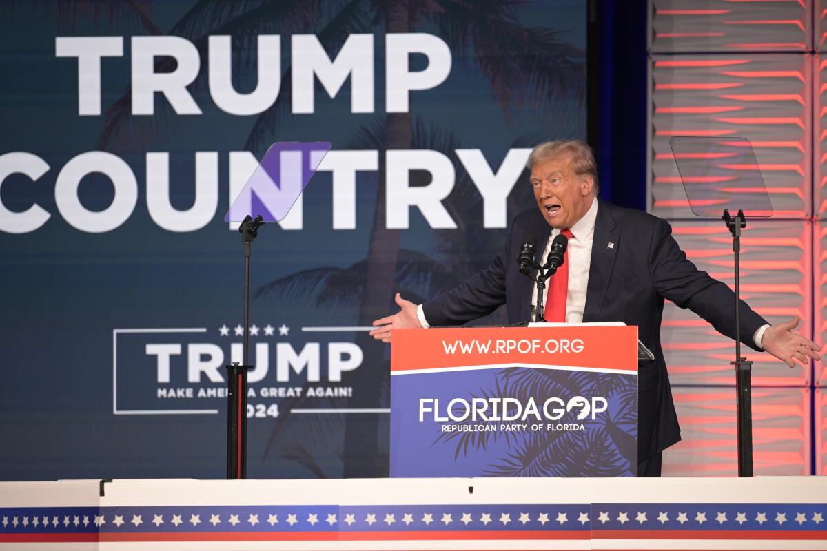 Former President Trump speaks with his arms outstretched in front of a sign reading "Trump Country."