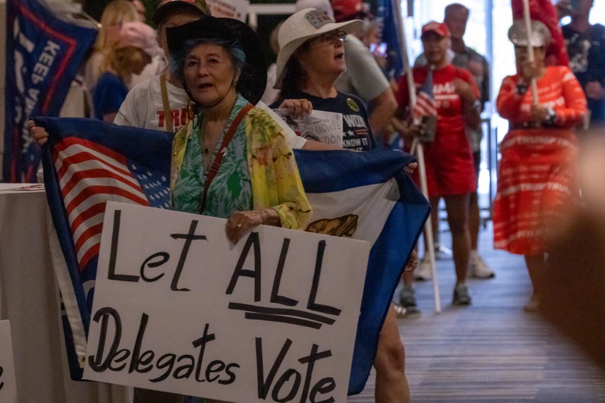 A group of demonstrators with Trump shirts, hats and banners, one holding a sign reading, "Let all delegates vote."