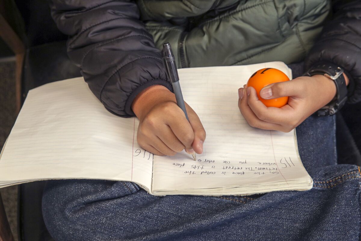 A child writes in a notebook with one hand while holding an orange stress ball in the other.