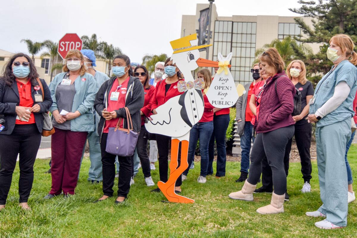 People in masks stand outside with a large sign shaped like a stork holding a bundle saying "Keep us open! Keep us public!"