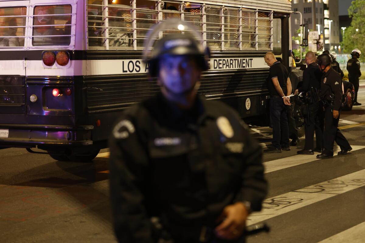 A police officer looks straight ahead while other officers arrest someone and load them on to a police bus