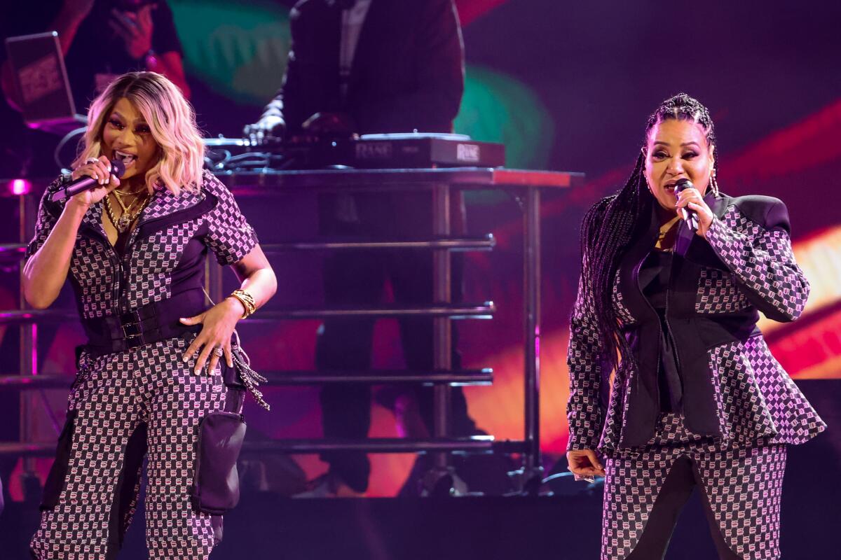 Two women in patterned outfits sing into microphones onstage.