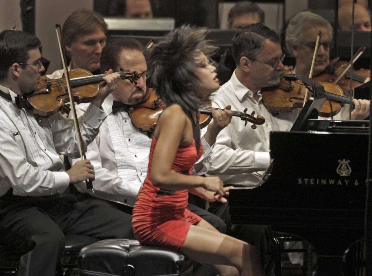 A woman in a red dress performs at the piano. Behind her violinists play.