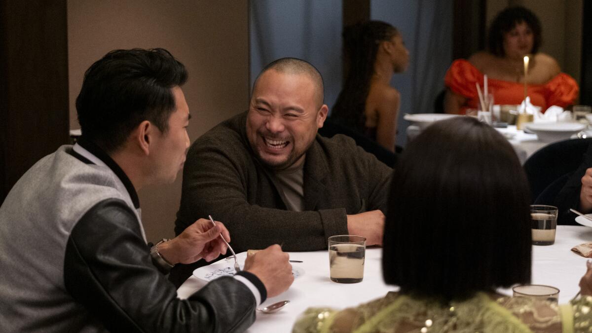 David Chang sitting at a dining table, smiling at the man to his right.