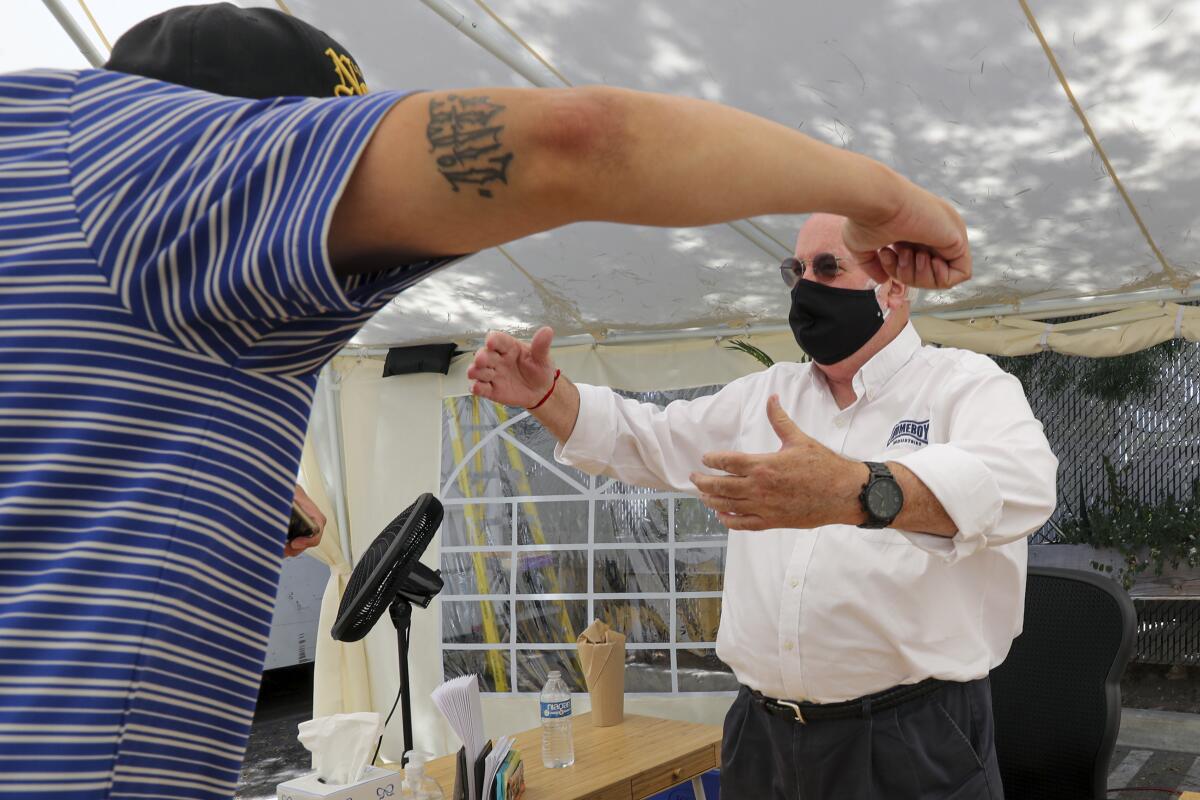 Father Greg Boyle, right, of Homeboy Industries, gives an air hug to a visitor under his tent office in the parking lot. 