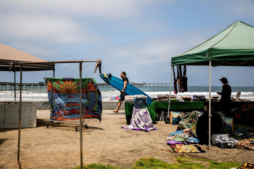 SAN DIEGO, CA - JULY 09: A surfer walk past street vendors in Ocean Beach on Friday, July 9, 2021 in San Diego, CA. (Sam Hodgson / The San Diego Union-Tribune)