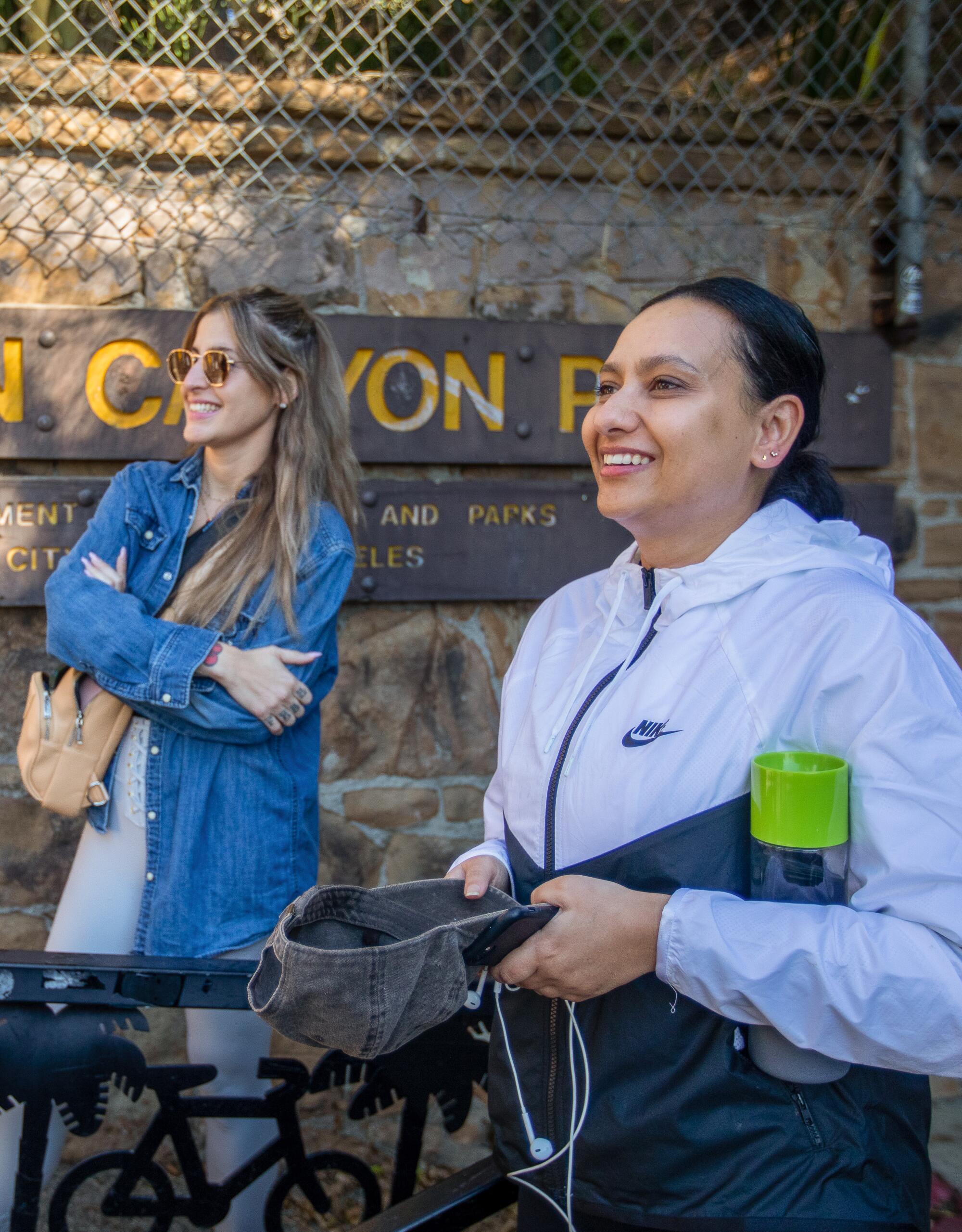 Two women in athletic clothing stand talking with others off-camera in front of a sign for Runyon Canyon Park.