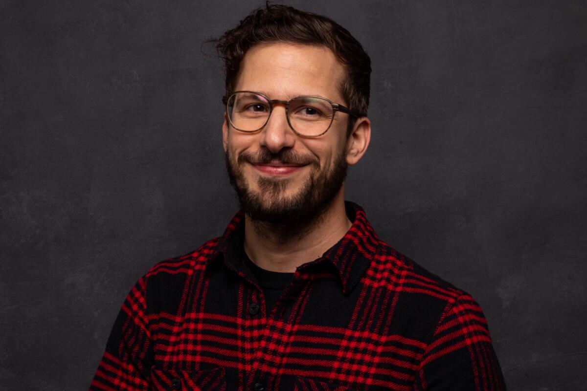 Andy Samberg smiling in glasses and a red-and-black plaid shirt in front of a dark background.