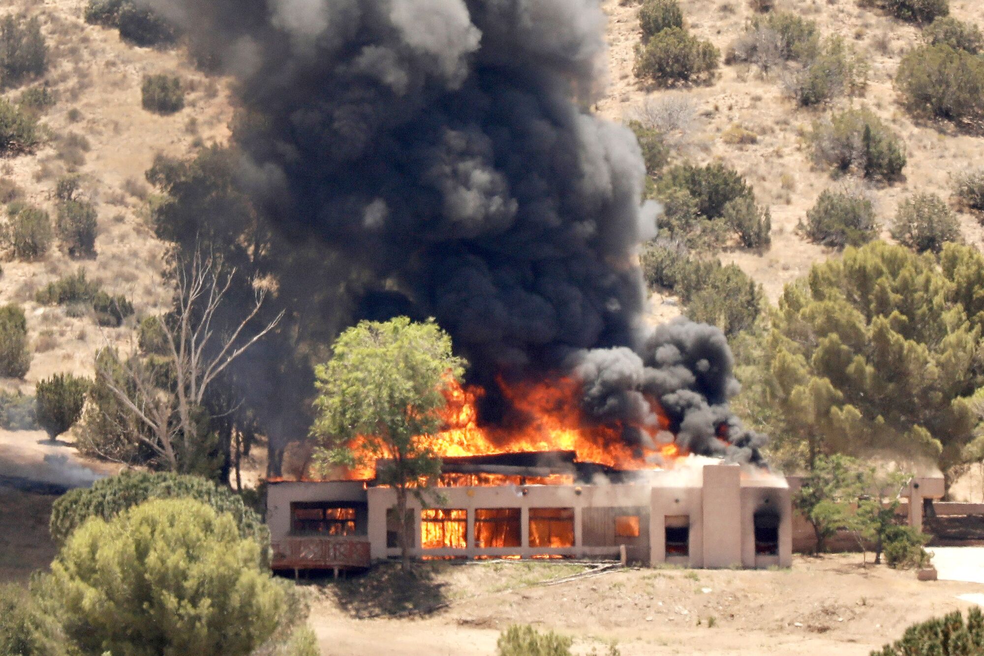 Bright orange flames fill a house on a sparsely vegetated hillside and send up a plume of black smoke.