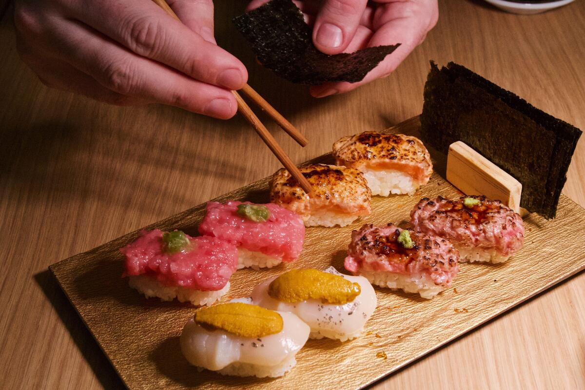 A hand holding chopsticks reaches for a selection of '"grab hand rolls" resembling nigiri, a nori sheet held in the other.