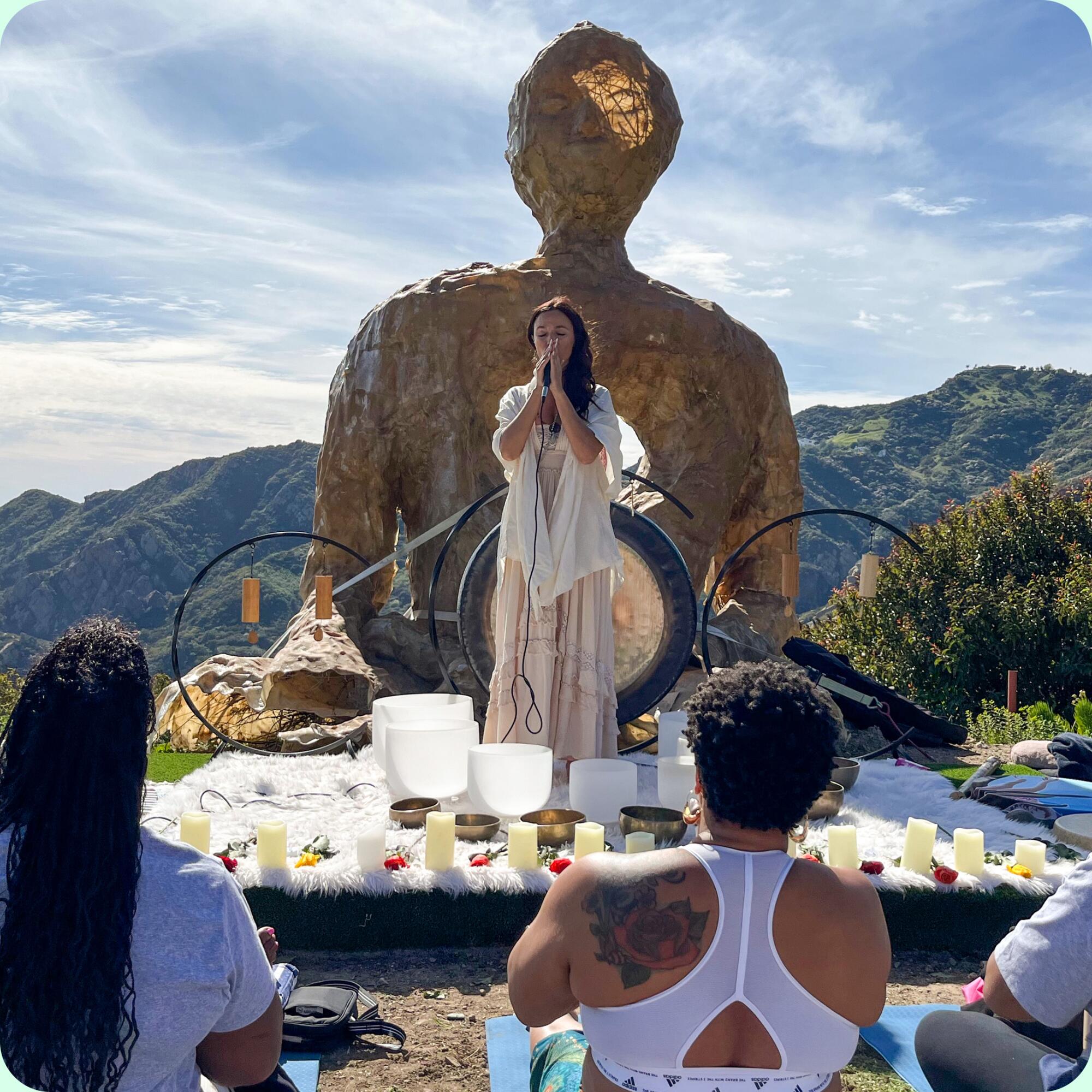 A woman stands on a shallow, candle-lined stage with her hands together. A large statue and a gong are seen behind her.