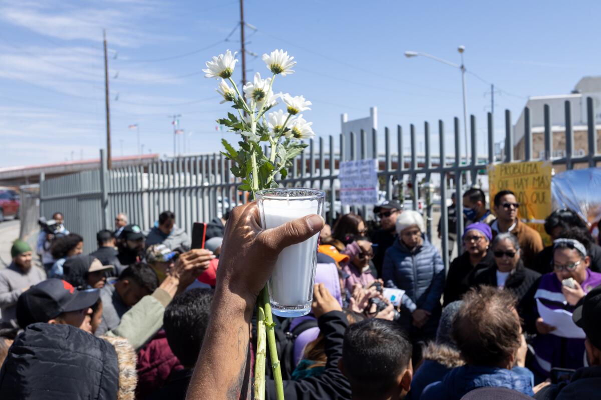 Migrants, one holding up flowers, at a fence around buildings in Ciudad Juárez, Mexico