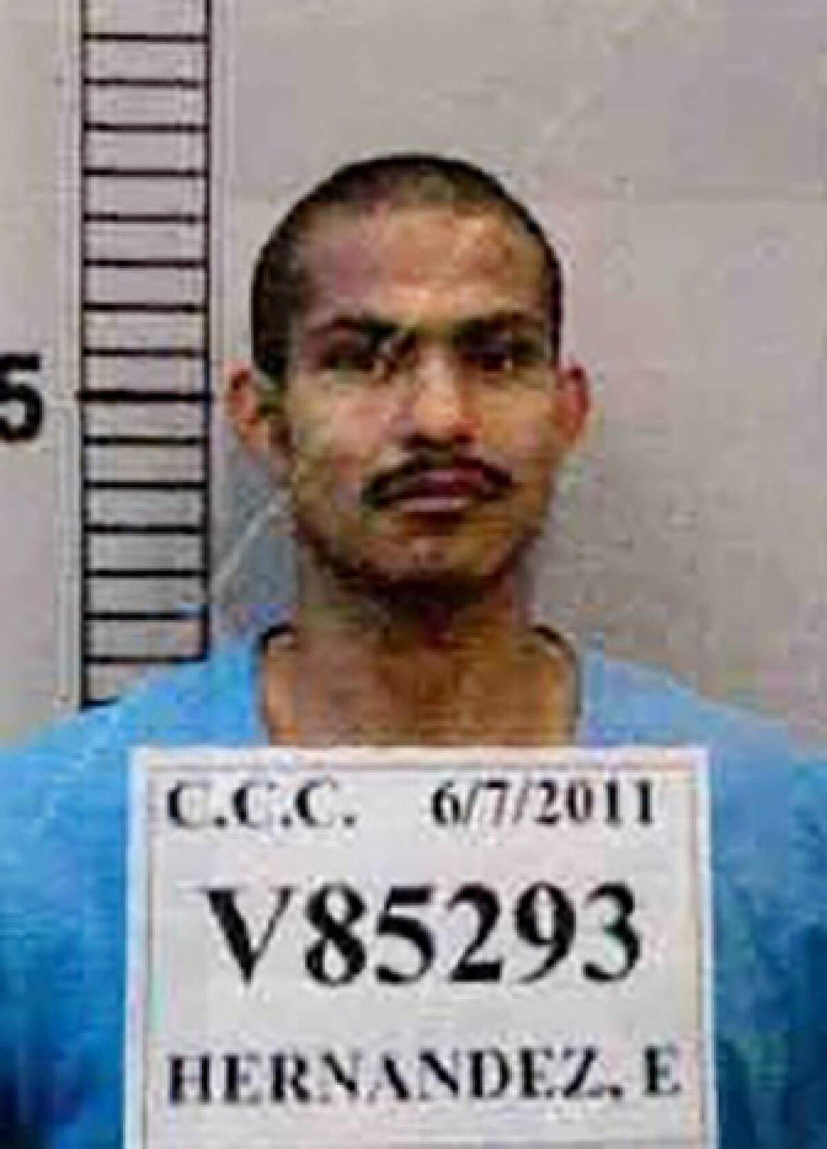 A booking photo of Eduardo Hernandez in blue jail garb, standing beside a height chart with a placard indicating arrest info