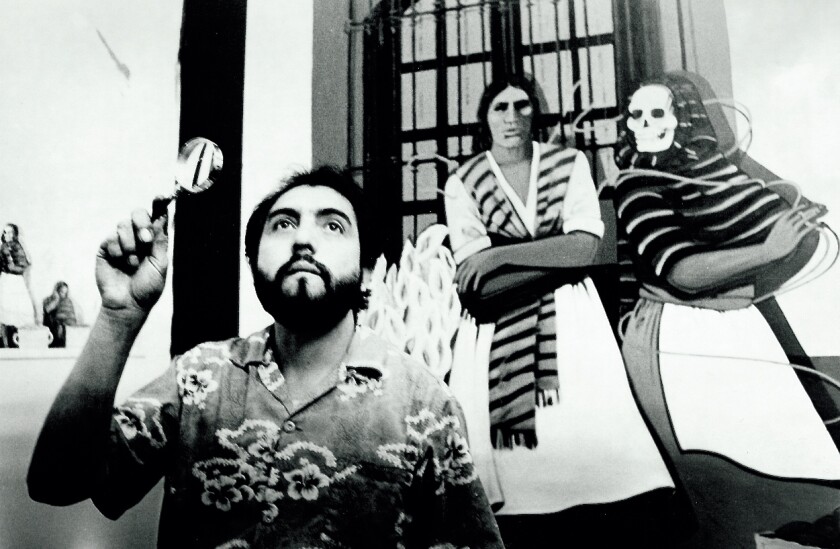 In a black-and-white photograph, Carlos Almaraz holds a magnifying glass while standing before two painted figures.