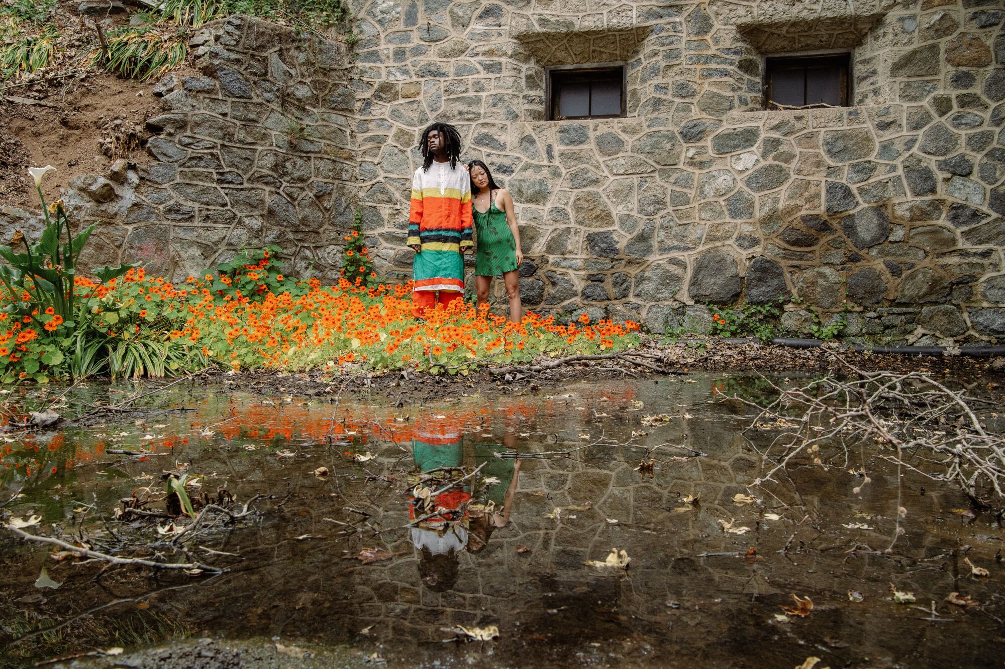 models pose in front of a stone structure among orange flowers with a swamp in front of them