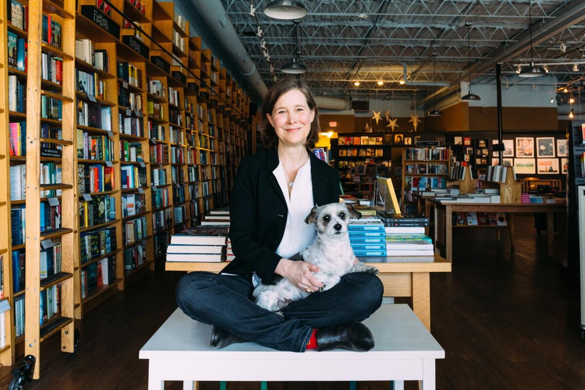 Author Ann Patchett sits on a table in a bookstore with a small dog on her lap.