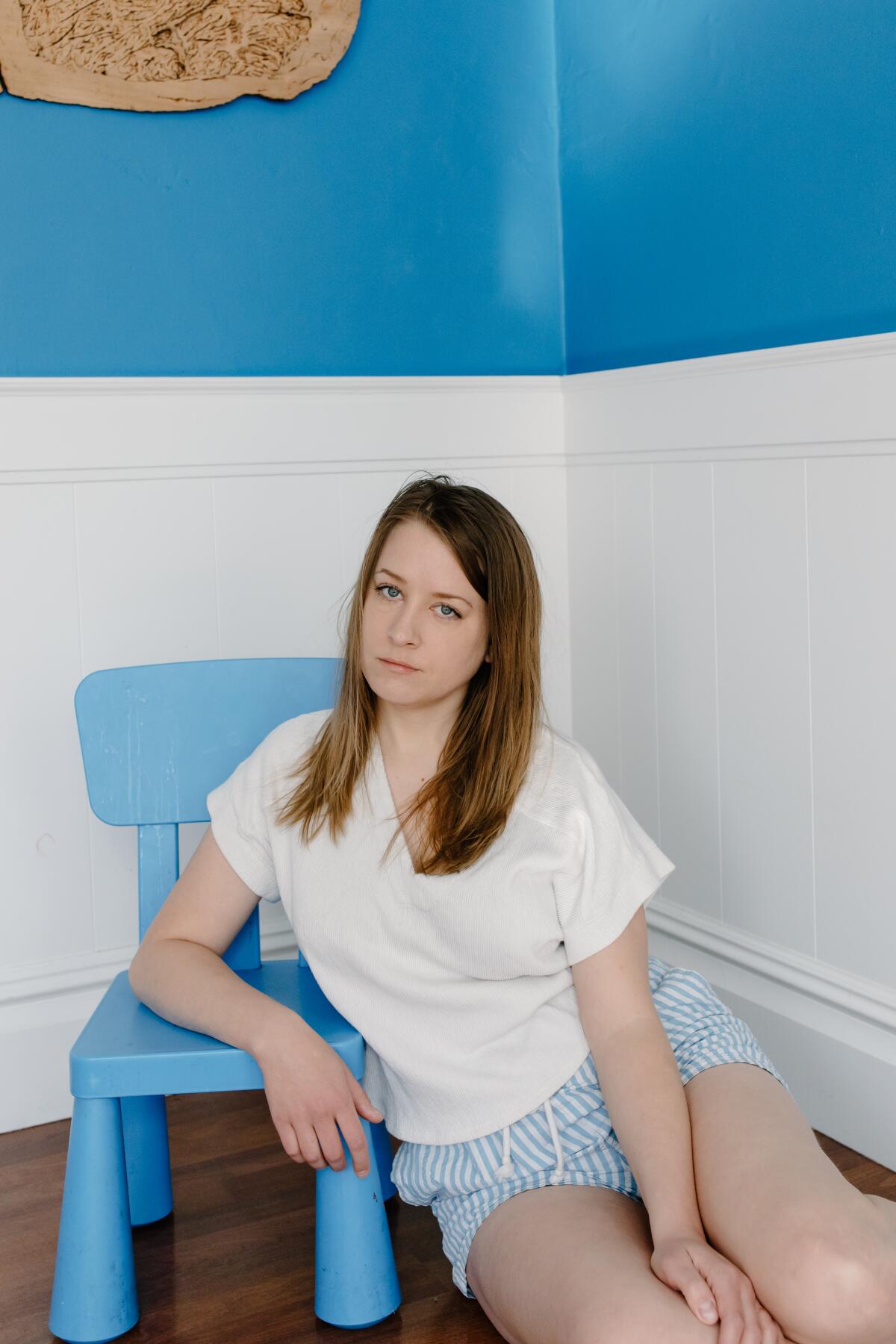 A woman in striped shorts and a white top leans against a chair in the corner of a room.