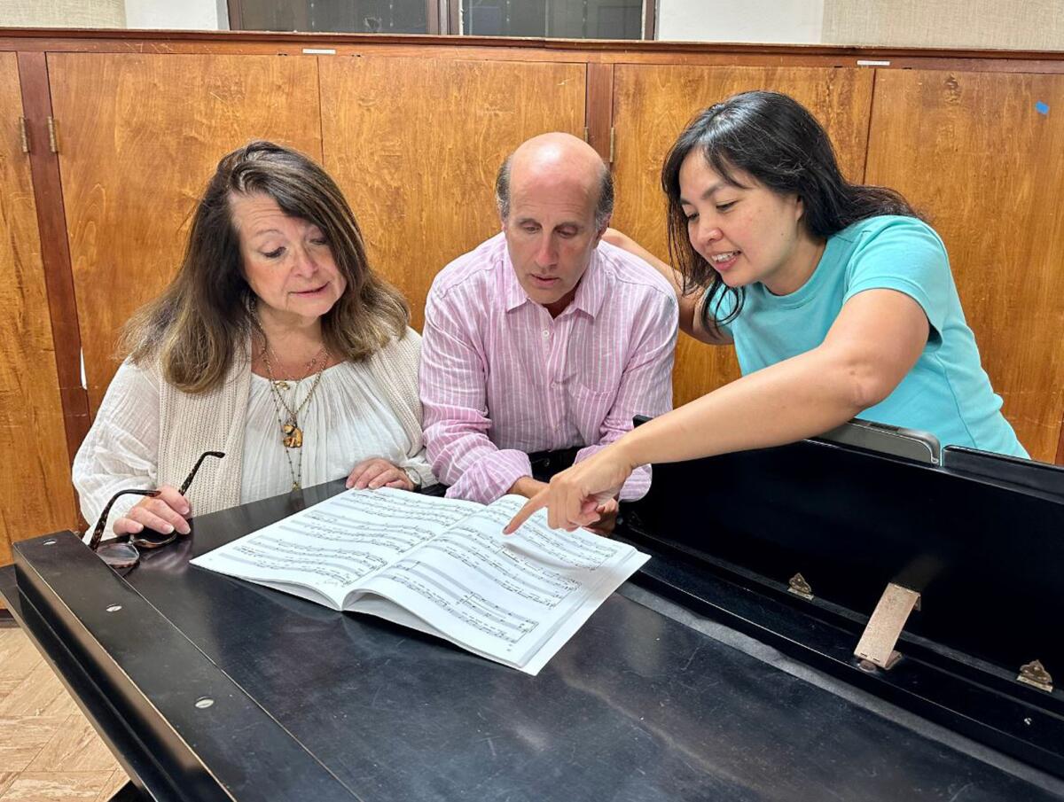 Three people look at sheet music around a piano.