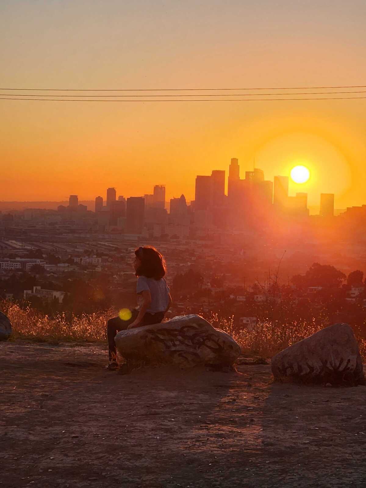 A little girl sits on the edge of a rock with the sunset and L.A. skyline in the background