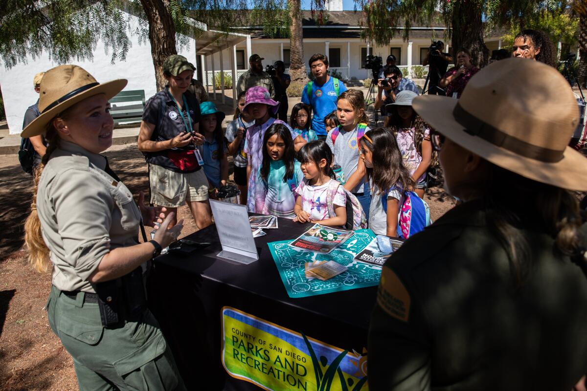 Two women in uniform speak to a group of children in the shade of large trees
