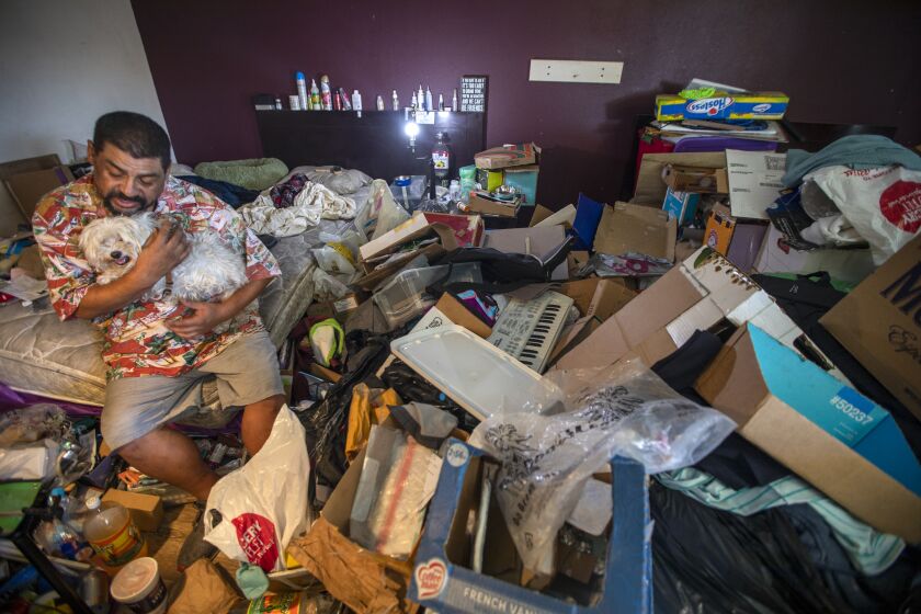 Downey, CA - June 30: Mario Blanco, 53, sat at the edge of his bed with his dog, "Leo the Lion," at Chateau Inn & Suites on Thursday, June 30, 2022, in Downey, CA. Blanco stares at the massive pile of cardboard boxes, clothes, and random items he's been hoarding in his hotel room for a year. (Francine Orr / Los Angeles Times)