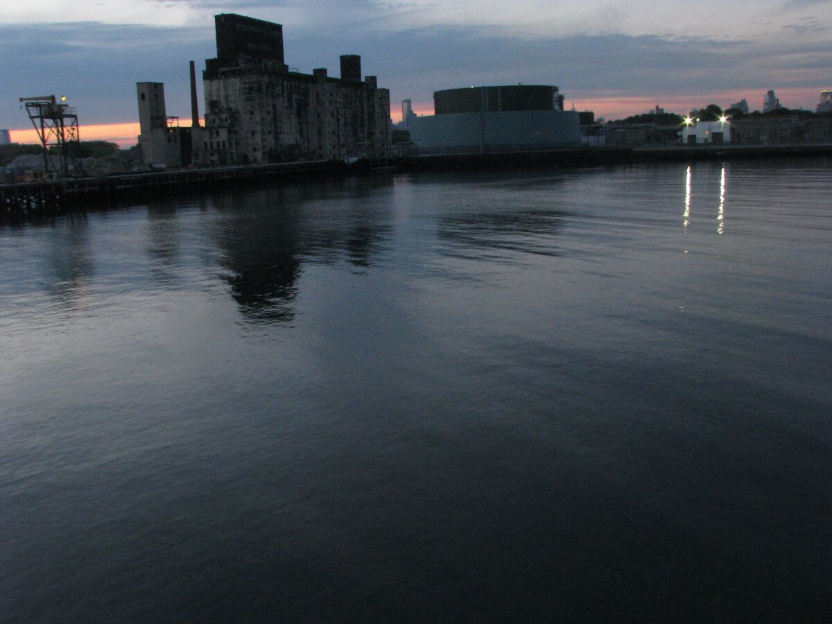A view of the Brooklyn waterfront at dusk shows industrial buildings in silhouette and the waters of New York Bay